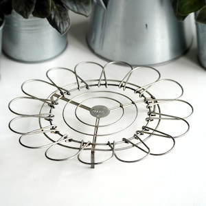 May include: A vintage metal trivet with a flower-like design. The trivet is made of silver-coloured wire and features a circular centre with radiating loops. The centre of the trivet has the word "MARS" stamped on it. The trivet is designed to protect surfaces from hot items.