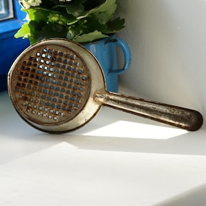 Pode incluir: Um passador de metal vintage com um padrão de grade quadrada e uma alça longa. O passador está ligeiramente enferrujado e tem uma aparência desgastada e antiga.