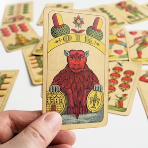 Puede incluir: Antigua carta de juego con una criatura roja con rostro humano, sosteniendo dos medallones dorados. La carta tiene la palabra "OTK" en una pancarta amarilla, con otras cartas visibles en el fondo. La carta se sostiene en una mano.