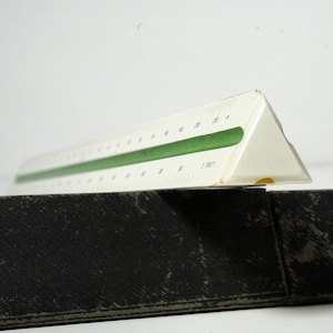 May include: A white and green triangular architect's scale ruler. The ruler has measurements in inches and is marked with white numbers and lines. The ruler is resting on a black surface.