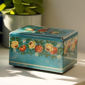 Pode incluir: Uma caixa de armazenamento retangular de metal azul com um padrão floral pintado de flores amarelas, laranjas e vermelhas com folhas verdes. A tampa exibe uma pequena ilustração de duas figuras e a caixa tem uma borda de linha branca.