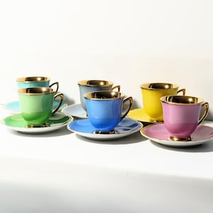 May include: A collection of six pastel-coloured teacups and saucers. Each cup and saucer is a different colour, including green, blue, yellow, and pink. The cups have gold rims and handles, and the saucers have gold trim.