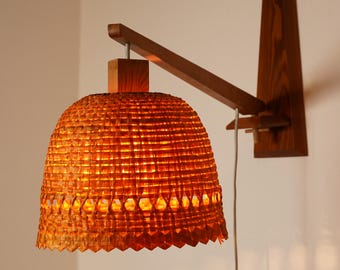 May include: A mid-century modern wall sconce with a woven orange lampshade. The lamp is made of wood and features a geometric design. The lampshade is a warm orange color, and the light shines through the woven pattern.