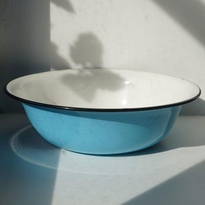 Pode incluir: Uma tigela redonda de esmalte com um exterior azul-celeste brilhante e um interior branco liso. A tigela tem uma borda preta fina e distinta que contorna a parte superior, proporcionando um contraste nítido com as superfícies do objeto.