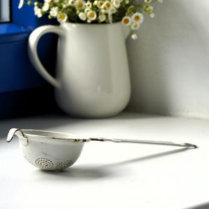 May include: A white enamelled metal tea strainer with a long handle and a perforated bowl featuring visible rust along the rim and base. The strainer is positioned horizontally, showcasing its simple construction and worn, vintage-style finish.