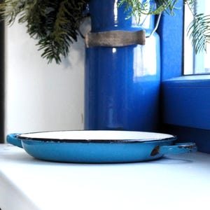 May include: A vintage, light blue enamel pan with two handles and a white interior. The pan is set on a white surface, with a cobalt blue vase and greenery in the background. The pan's rim has a black trim.