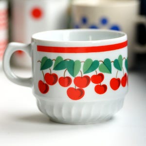 Può includere: Tazza da tè in ceramica bianca con una fascia rossa e un motivo a ciliegie. Le ciliegie sono rosse con foglie verdi.