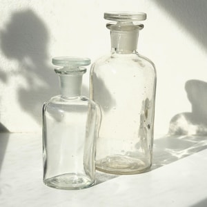 Könnte beinhalten: Zwei durchsichtige Apothekerflaschen aus Glas mit passenden Glasstopfen. Die Flaschen haben einen zylindrischen Körper mit abgerundeter Schulter und einem kurzen Hals. Das Glas ist transparent und glatt und zeigt ein schlichtes Design.