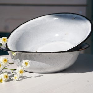 May include: A white enamel bowl with a speckled finish and a dark rim. The bowl has a wide, shallow basin and a smooth, glossy surface. A small cluster of white flowers with yellow centres is positioned in the foreground, partially overlapping the base.