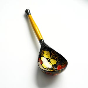 Peut inclure: Une cuillère en bois avec un long manche jaune et un bol noir. Le bol est orné d'un motif floral doré et d'accents rouges. La cuillère est probablement utilisée pour servir ou cuisiner.