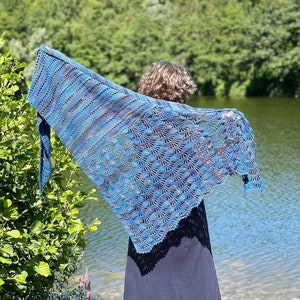 Op de afbeelding: Een persoon draagt ​​een blauwe en paarse gehaakte sjaal met een gegolfde rand. De sjaal is over de schouders en rug van de persoon gedrapeerd. De persoon staat voor een waterpartij met groene bomen op de achtergrond.