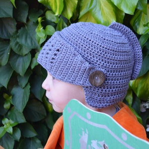 May include: A child wearing a gray crocheted hat with a visor and a button. The hat has a unique design resembling a helmet. The child is wearing an orange shirt and holding a green shield.