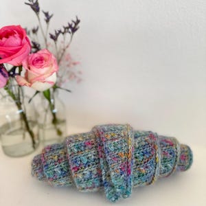 May include: A handmade, crocheted croissant in shades of blue, green, and pink, with a textured appearance. The pastry is displayed on a white surface, with a vase of pink roses and purple flowers in the background.