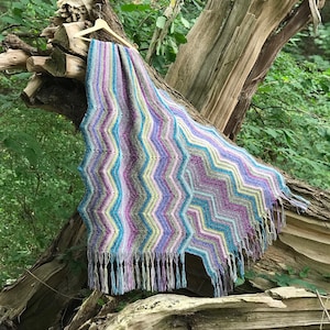 Op de afbeelding: Een veelkleurig gehaakt deken met een chevron-patroon en franjes die aan een tak in een bos hangt.