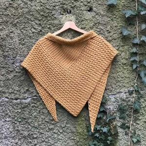 Peut inclure: Un châle en crochet jaune moutarde suspendu à un cintre en bois contre un mur de pierre texturé avec du lierre vert qui pousse dessus.