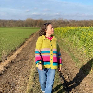 Op de afbeelding: Een persoon draagt een gele, roze, blauwe en groene gestreepte gehaakte cardigan met blauwe jeans en zwarte schoenen en loopt over een onverhard pad.