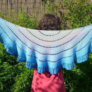 May include: A blue and white crocheted shawl with a scalloped edge. The shawl is being held by a person in a garden setting.