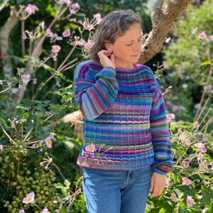 Puede incluir: Una mujer lleva un suéter de rayas de colores con un patrón texturizado. El suéter está hecho de un hilo suave y tiene un ajuste holgado. La mujer está de pie en un jardín con flores rosas.