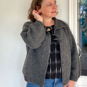 Op de afbeelding: Een grijze gebreide cardigan met een kraag en knoopsluiting. De cardigan wordt gedragen over een zwart-wit geblokt shirt en een blauwe jeans.