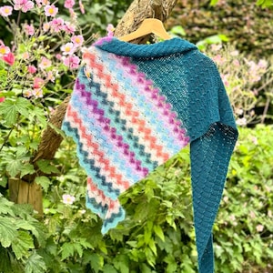 Op de afbeelding: Een teal en veelkleurige gehaakte sjaal met een gegolfde rand hangt aan een houten hanger voor een groene, bladerrijke achtergrond.