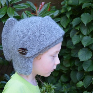 Puede incluir: Un gorro de punto gris con orejeras y un botón decorativo. El gorro lo lleva puesto un niño.