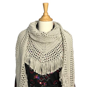 Peut inclure: Un châle en crochet avec une bordure à franges de couleur beige clair. Le châle est drapé sur un mannequin.