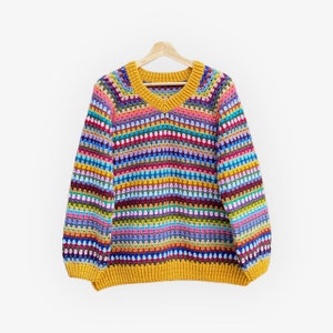 Puede incluir: Un suéter de ganchillo hecho a mano, colorido, con cuello en V y mangas largas. El suéter presenta rayas horizontales en varios colores, incluyendo amarillo, rosa, azul y verde. El cuello y los puños son de color amarillo mostaza liso. El suéter está colgado en una percha de madera.