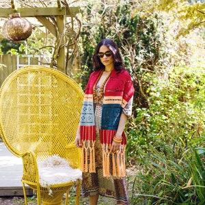 May include: A woman wearing sunglasses and a colorful crocheted shawl with fringe, standing in front of a yellow wicker chair.