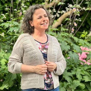 May include: A woman wearing a beige crocheted cardigan with a floral print shirt underneath. She is standing in a garden with green foliage and pink flowers.
