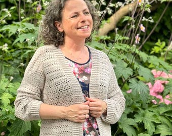 Puede incluir: Una mujer lleva una chaqueta de punto de ganchillo beige con una camisa de estampado floral debajo. Está de pie en un jardín con follaje verde y flores rosas.