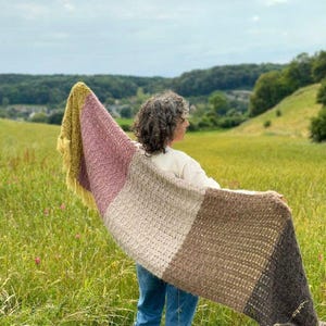 May include: A person wearing blue jeans and a white shirt holds a multi-colored crocheted shawl in a field.