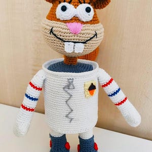 Puede incluir: Un juguete de peluche de ganchillo de una ardilla de dibujos animados que lleva un traje de astronauta blanco con rayas rojas y azules en las mangas. La ardilla tiene un cuerpo marrón, una cara blanca y grandes ojos negros.