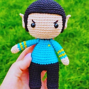 May include: A crocheted toy figure of a person with black hair, a blue shirt, and black pants. The toy has a serious expression with furrowed brows and large eyes.