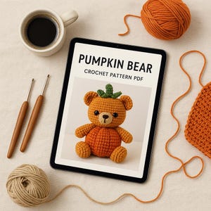 May include: A tablet shows a crochet pattern for a Pumpkin Bear. The bear is orange with a green stem. Two crochet hooks, a cup of coffee, and yarn are also in the image.