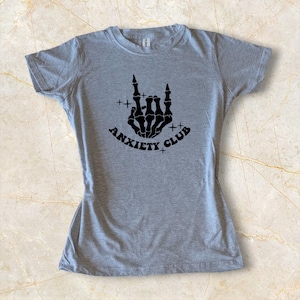 May include: A gray t-shirt featuring a black graphic of a skeleton hand making a rock and roll gesture, with the words "ANXIETY CLUB" in a curved font below. The shirt is made of a soft material.