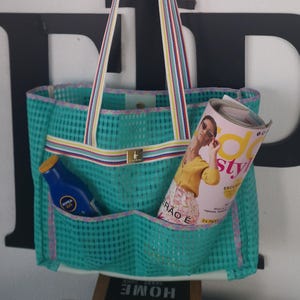 Könnte beinhalten: Eine türkisfarbene Netz-Tragetasche mit bunten, gestreiften Trägern, die an einem Haken hängt. Die Tasche enthält ein Magazin und eine blaue Flasche. Das Magazincover zeigt eine Frau mit Sonnenbrille und einem gelben Oberteil mit dem Text "Style" auf dem Cover.