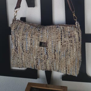 Könnte beinhalten: Eine braune gewebte Umhängetasche mit einem braunen Riemen, der an einem Haken hängt. Die Tasche hat ein gewebtes Muster mit blauen und braunen Fäden.