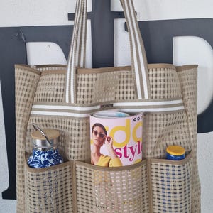 Könnte beinhalten: Eine beige und weiß gestreifte Tragetasche mit mehreren Taschen. Die Tasche ist gefüllt mit einem Magazin, einer Wasserflasche und einer kleinen Flasche Sonnencreme. Das Magazincover zeigt eine Frau mit Sonnenbrille und einem gelben Oberteil mit dem Text "add style".