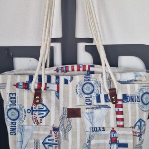 May include: A white canvas tote bag with a nautical theme. The bag features red and blue stripes, lighthouses, anchors, and the text "Nautical North Coast Est. 1924" and "Never Stop Exploring".