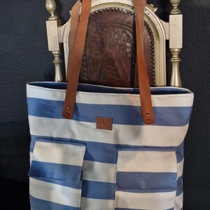May include: A blue and white striped canvas tote bag with brown leather straps and two large pockets on the front. The bag has a small, white square label with a logo on the front.
