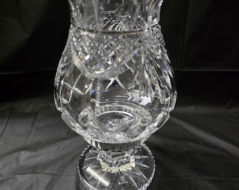 May include: A clear, cut-glass vase with a flared top and a pedestal base. The vase features intricate diamond and starburst patterns, reflecting light. The design includes a scalloped rim and a detailed, textured surface, suitable for home decor.