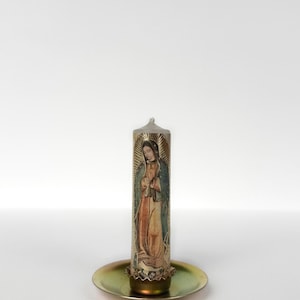 Op de afbeelding: Een cilindrische kaars met een afbeelding van een religieuze figuur, geplaatst op een messingkleurige metalen plaat. De kaars toont een figuur in een gewaad met een halo, tegen een zilveren achtergrond.