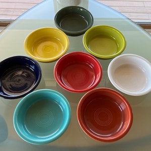May include: A collection of eight round ceramic bowls in various solid colors including dark green, yellow, lime green, navy blue, red, white, turquoise, and orange. Each bowl features a smooth interior surface and a distinct concentric ring pattern.