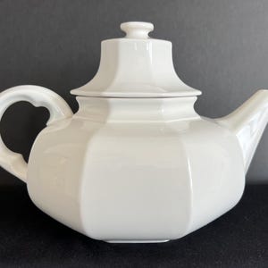 May include: A white ceramic teapot with an octagonal body, a matching lid with a small knob handle, a curved spout, and a C-shaped handle. The teapot features clean, geometric lines and a smooth, glossy finish, presenting a solid white color throughout.