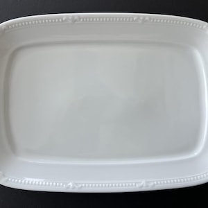 May include: A rectangular white ceramic serving platter with a slightly raised rim and rounded corners. The rim is decorated with a delicate beaded pattern and small floral accents. The platter is set against a black background.