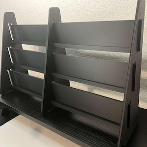 May include: A dark gray wooden magazine rack with multiple shelves. The rack has a modern design with a slatted back and angled sides. The shelves are evenly spaced and appear sturdy, suitable for organizing reading materials.