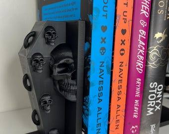 May include: A black coffin-shaped bookend with skull details supports several books. The books have colourful spines with titles like "Lights Out" and "Caught Up." The bookend is made of a dark material, adding a gothic touch to a bookshelf.