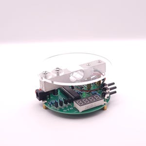 May include: A small, circular circuit board featuring a digital display and various electronic components. The board is green with silver and black elements, and a clear, circular cover sits on top. The display reads "3461BS-1".