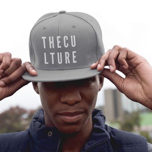 May include: A gray baseball cap with the text "THE CULTURE" in white block letters. The cap is being held by a person. The person is wearing a navy blue vest and a gray long-sleeve shirt.