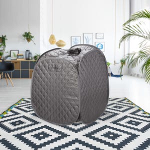 May include: A gray portable sauna tent with a quilted exterior. The tent is set up in a modern living room with a white rug and a potted plant.